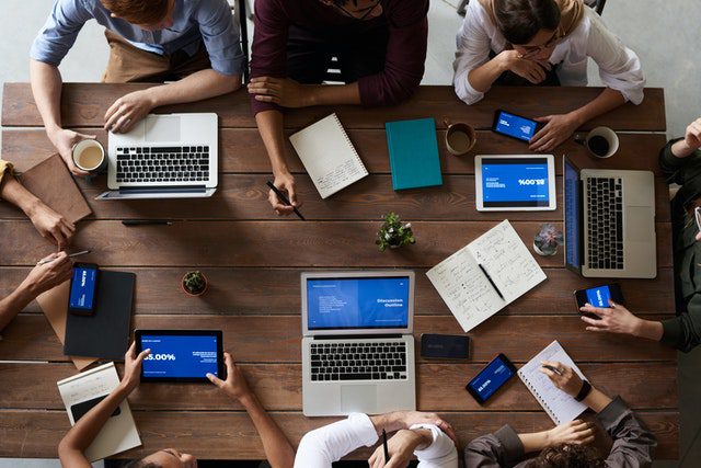 Equipe de développement web collaborant autour d'une grande table en bois, utilisant des ordinateurs portables, des tablettes et des smartphones pour travailler sur des projets.